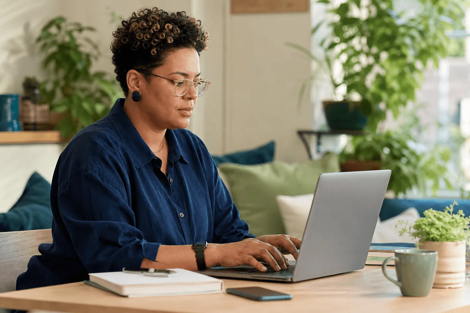 An adult typing on a laptop in a bright cosy space with phone and notebook nearby, focused and relaxed.